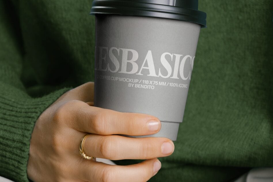Gray coffee cup with black lid held in hand wearing gold ring, green sweater background