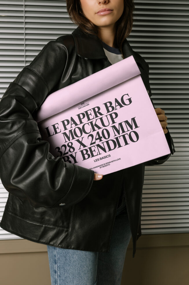 Woman holding pink paper bag mockup against metal shutter wall in black leather jacket