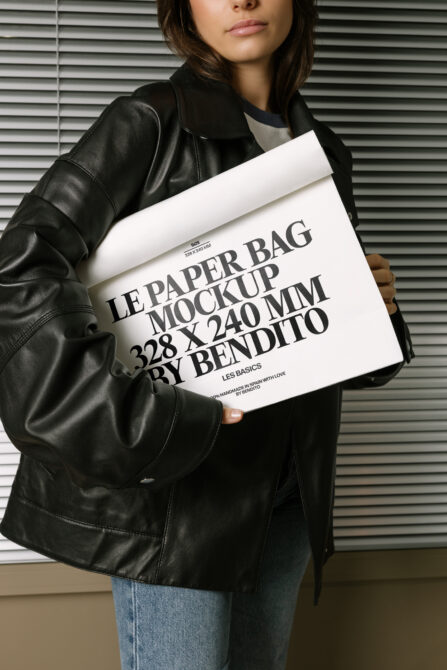 Woman holding white paper bag mockup against metal shutter background wearing black jacket