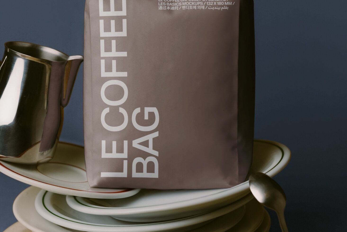 Styled tabletop with stacked white plates, metal pitcher, taupe coffee bag mockup on blue background