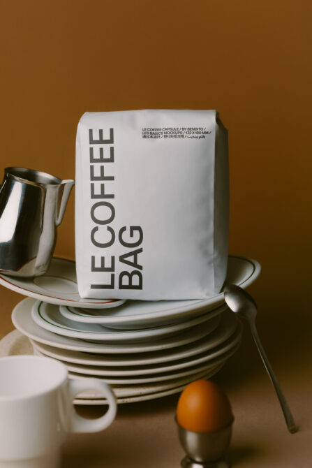 Styled tabletop with stacked white plates, metal pitcher, white coffee bag mockup on warm background