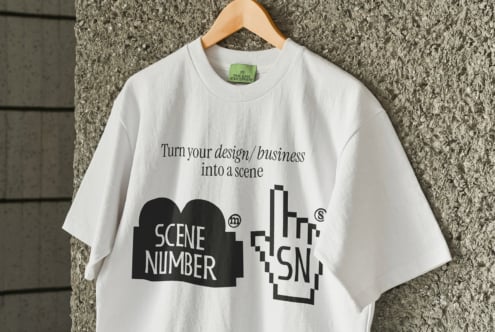 White tee with Scene Number branding on wooden hanger against concrete wall