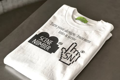 Cream colored t-shirt folded with Scene Number tea design on gray surface