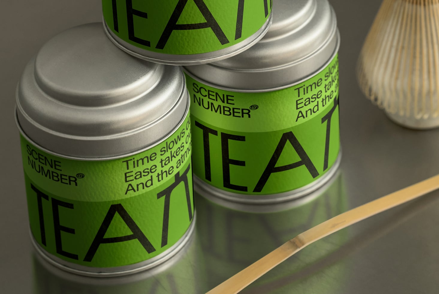 Two silver tea tins with green labels stacked on wooden surface with whisk visible