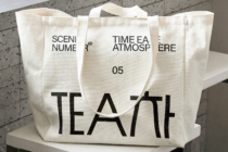 Natural canvas tote with bold black TEA text, displayed on platform, front view