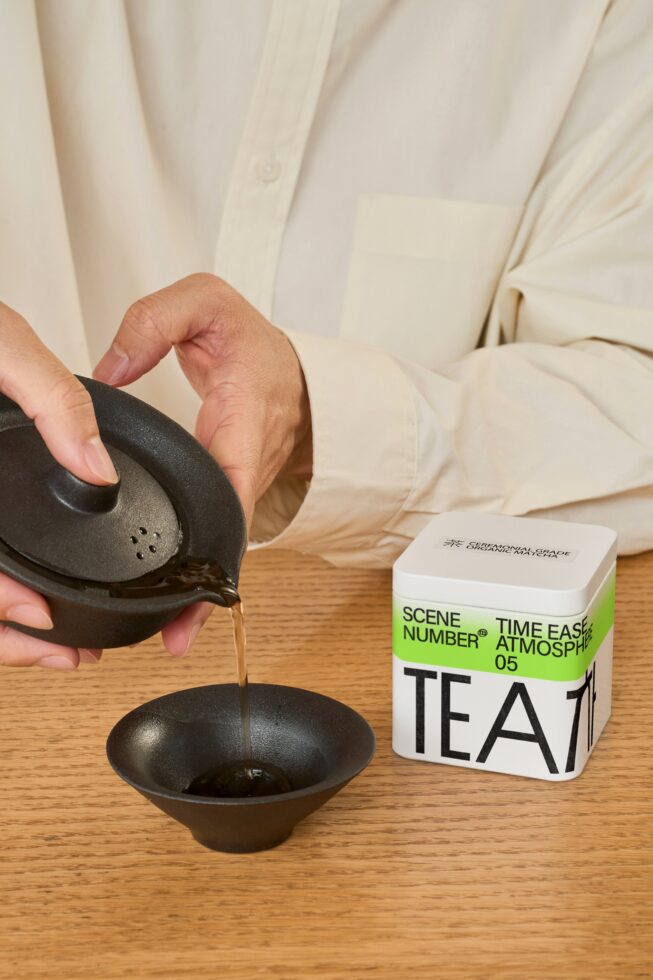 Pouring tea from black ceramic pot into white tea box next to wooden surface