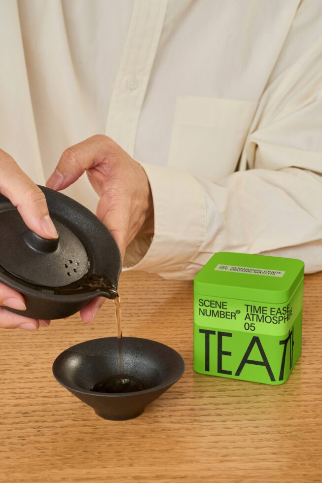 Pouring tea from black ceramic pot into bowl next to green tea packaging box