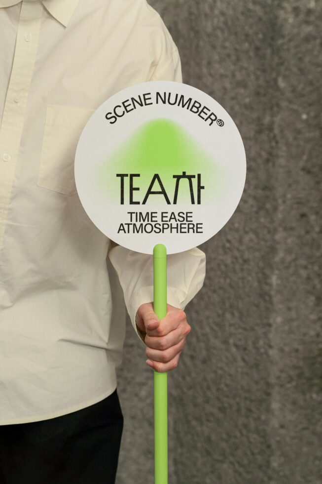 Round promotional sign with Scene Number branding held by person in cream shirt