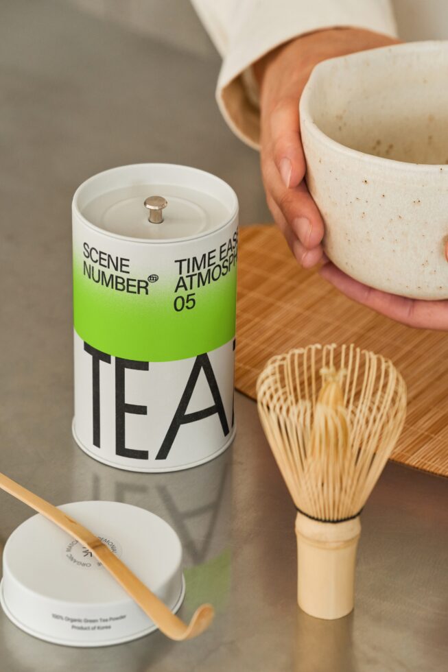 White tea candle with lime band label, matcha whisk nearby on wood surface
