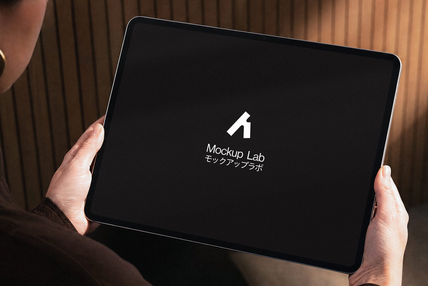 Hands holding tablet with Mockup Lab logo on dark screen, warm curtain background