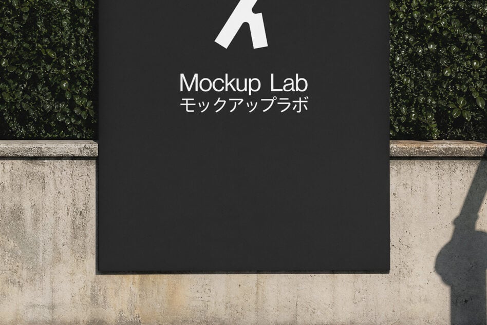 Outdoor storefront sign mockup with dark panel, white logo, bilingual text, concrete base, green hedges, natural shadow