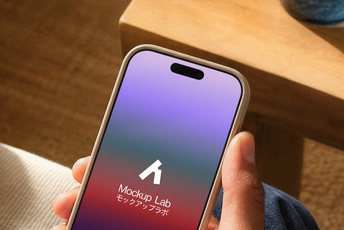 Hand holding beige iPhone with purple to red gradient screen displaying Mockup Lab logo on wooden desk