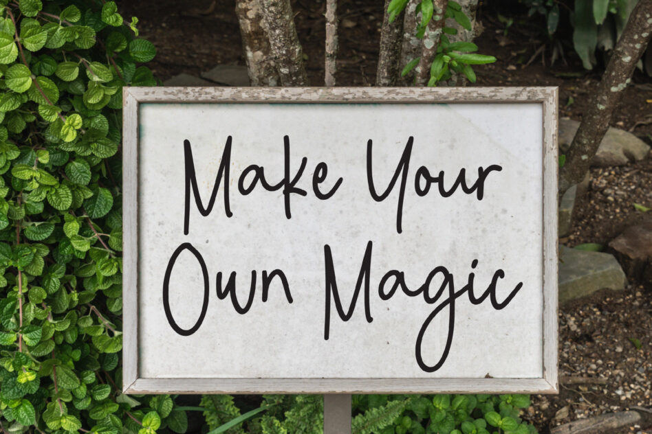 Sign with handwritten style font reads Make Your Own Magic surrounded by greenery, perfect for graphic design inspiration, fonts, nature-themed mockups.