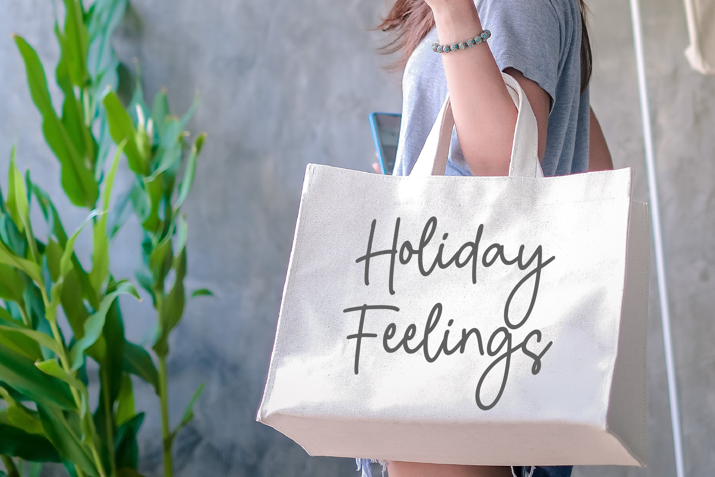 Woman in gray tee holding natural canvas tote bag with Holiday Feelings script text