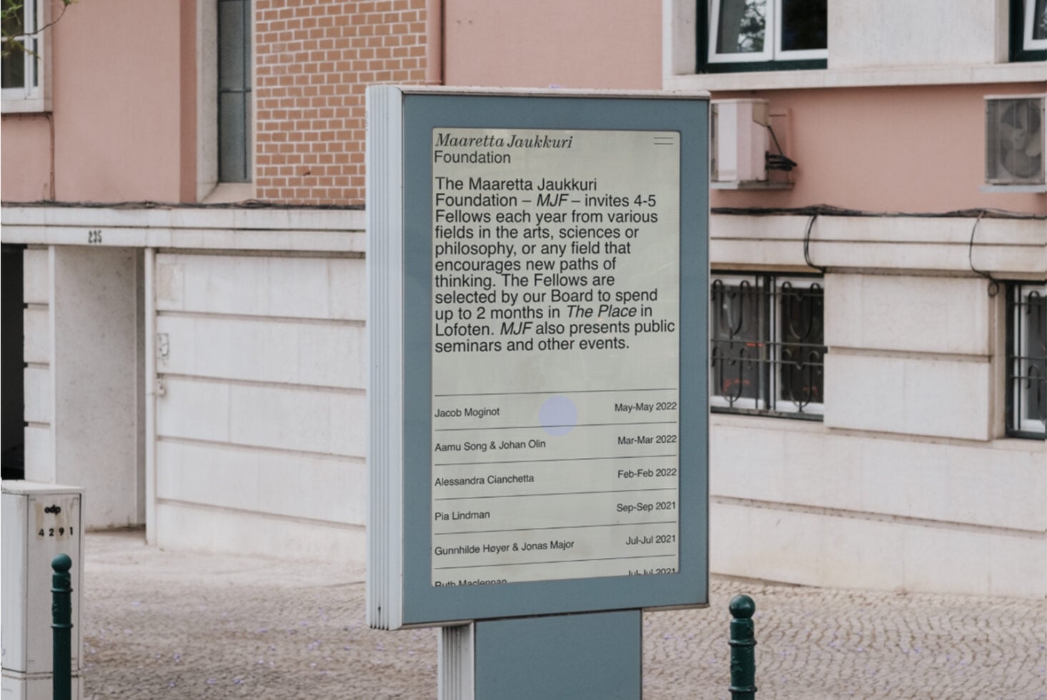 Outdoor wayfinding sign with cream panel listing Maaretta Jaukkuri Foundation fellows on blue-grey post in courtyard
