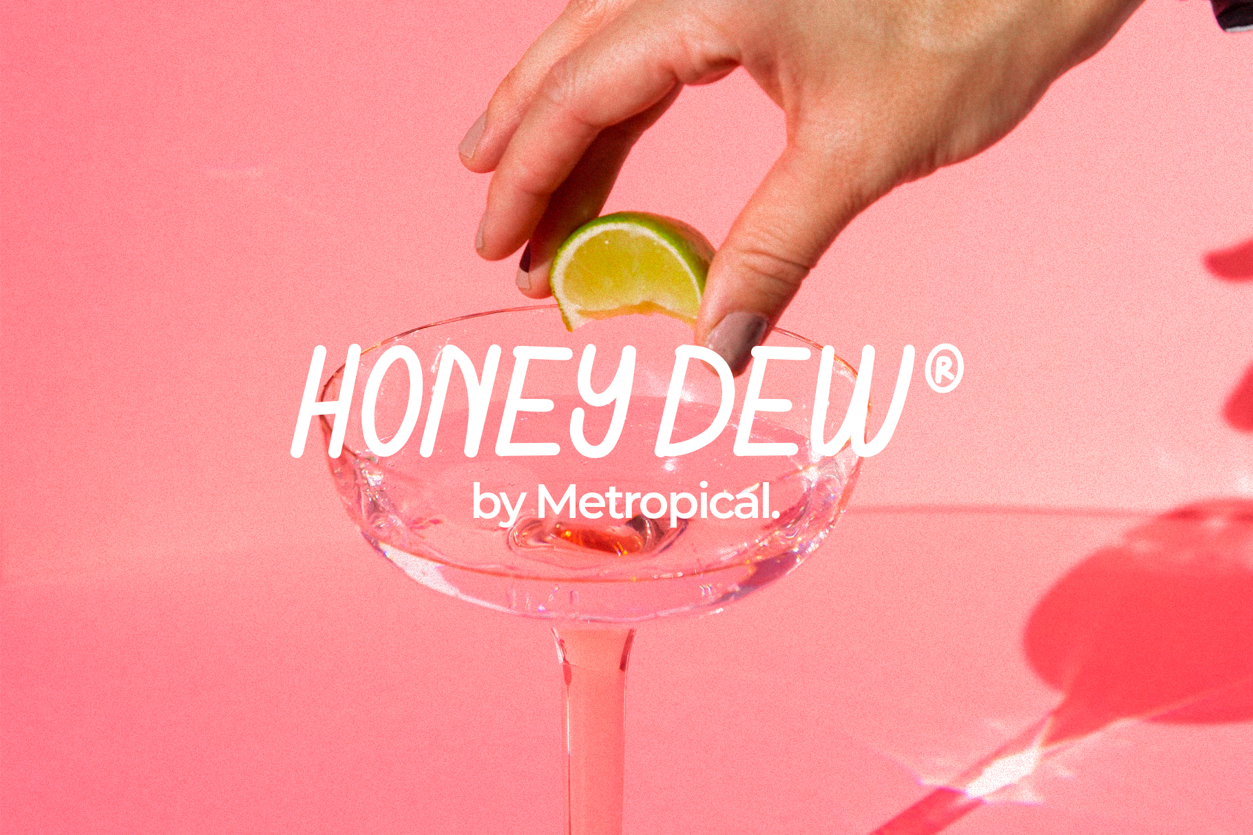 Hand dropping lime slice into pink cocktail with Honey Dew branding on coral background