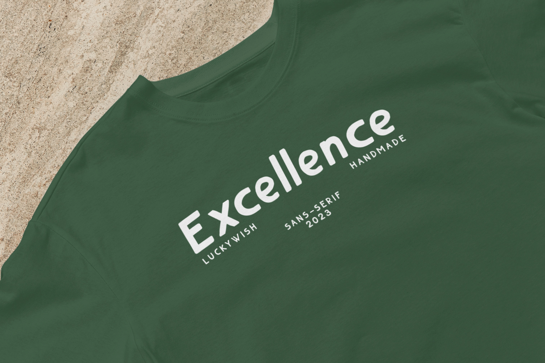 Green t-shirt with white Excellence Handmade logo printed on sandy surface backdrop
