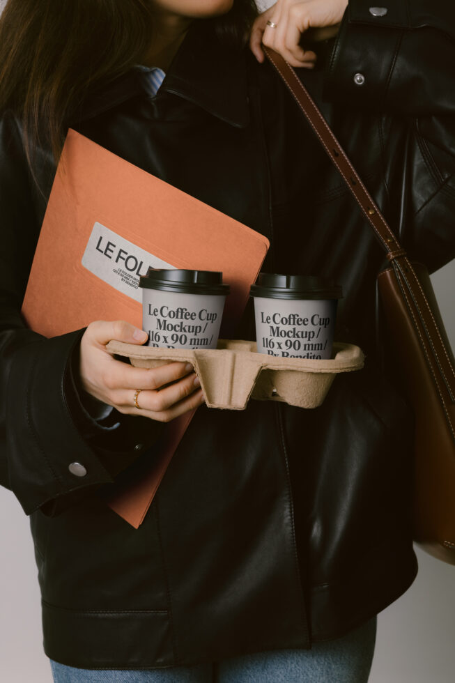 Woman in black leather jacket holding two coffee cups in cardboard carrier, orange folder visible