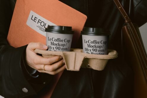 Close-up of hands holding two coffee cup mockups in carrier with orange folder background