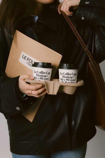 Woman in black leather jacket holding two coffee cups in cardboard carrier, beige folder visible