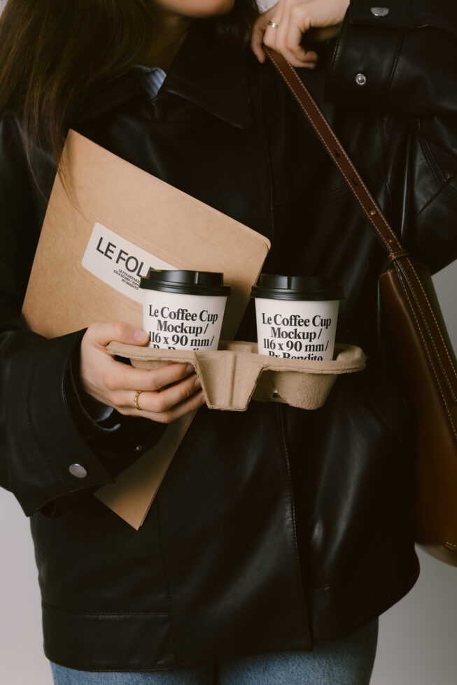 Woman in black leather jacket holding two coffee cups in cardboard carrier, beige folder visible