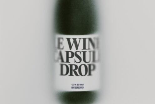 Close-up of skincare bottle label reading "SERUM CAPSULE DROP" with specs listed below, blurred green bottle behind