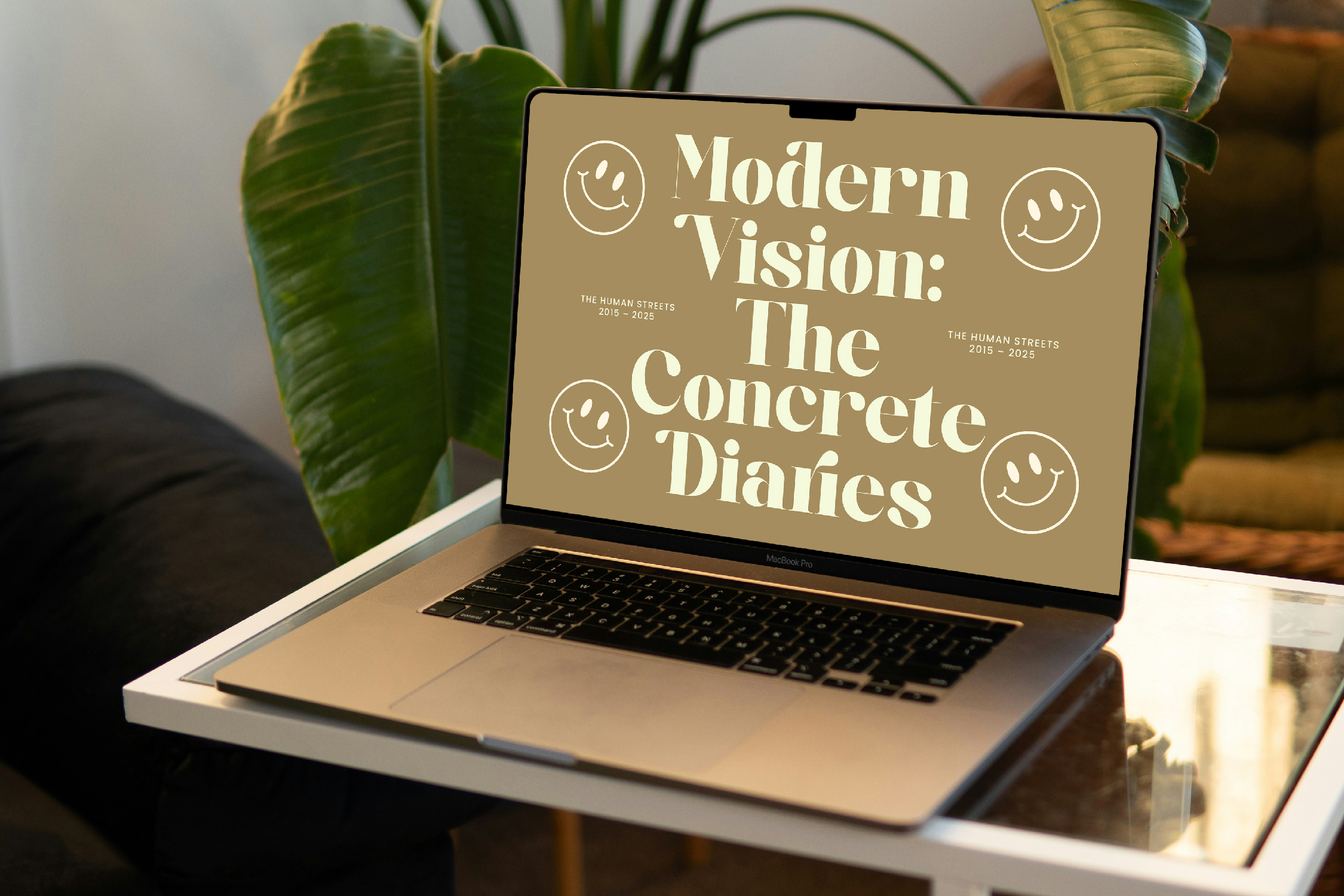 MacBook Pro displaying beige poster with "Modern Vision: The Concrete Diaries" title, smiley faces, surrounded by potted plants on desk