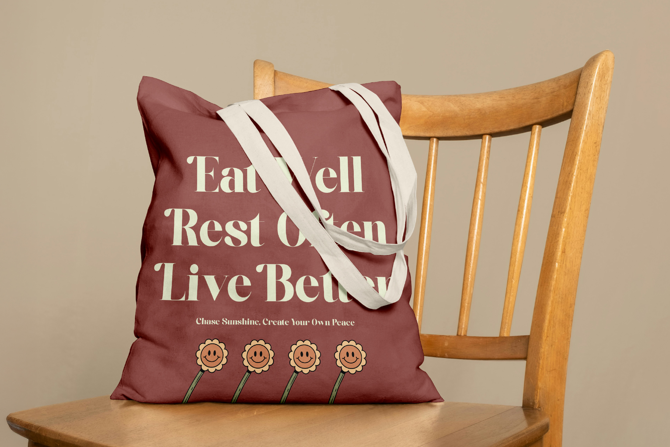 Burgundy tote bag with white text reading "Eat Well Rest Often Live Better" plus smiley flower illustrations, draped on wooden chair