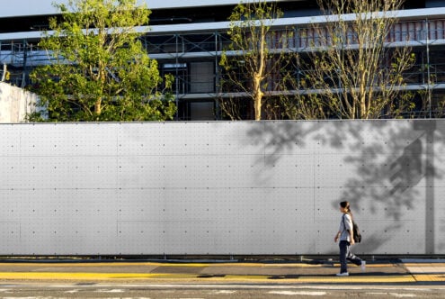 Woman walking past concrete wall with tree shadows in bright sunlight, urban architecture