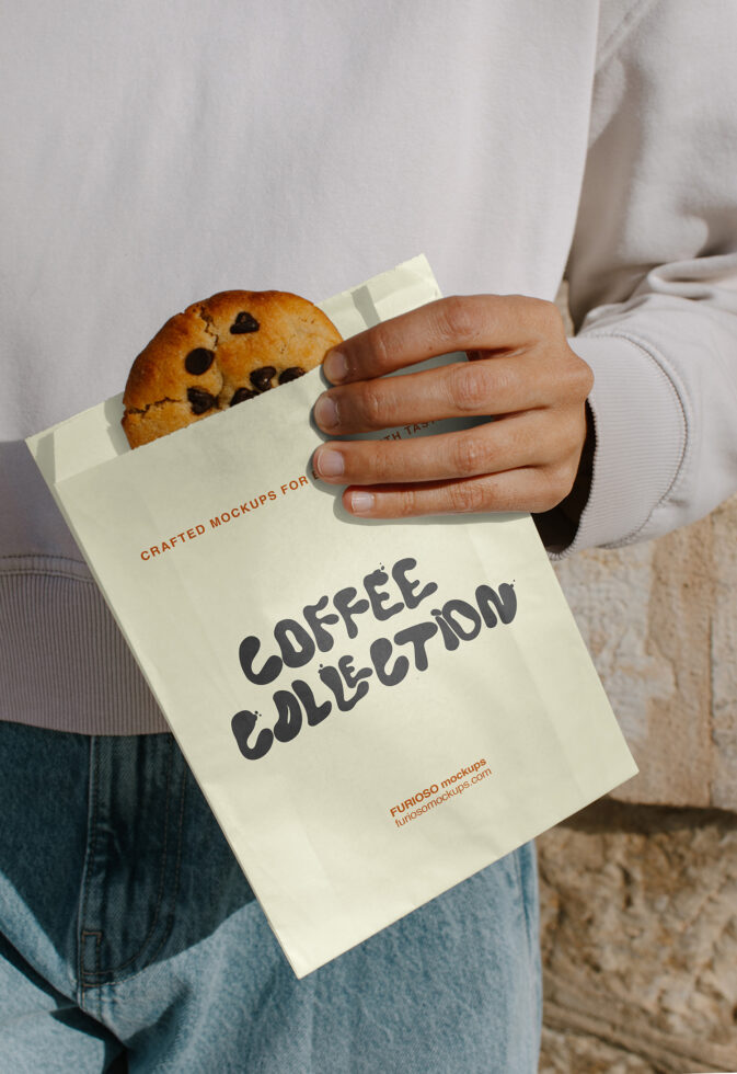 Cream paper bag with chocolate chip cookie peeking out, held in hands with dark nail polish