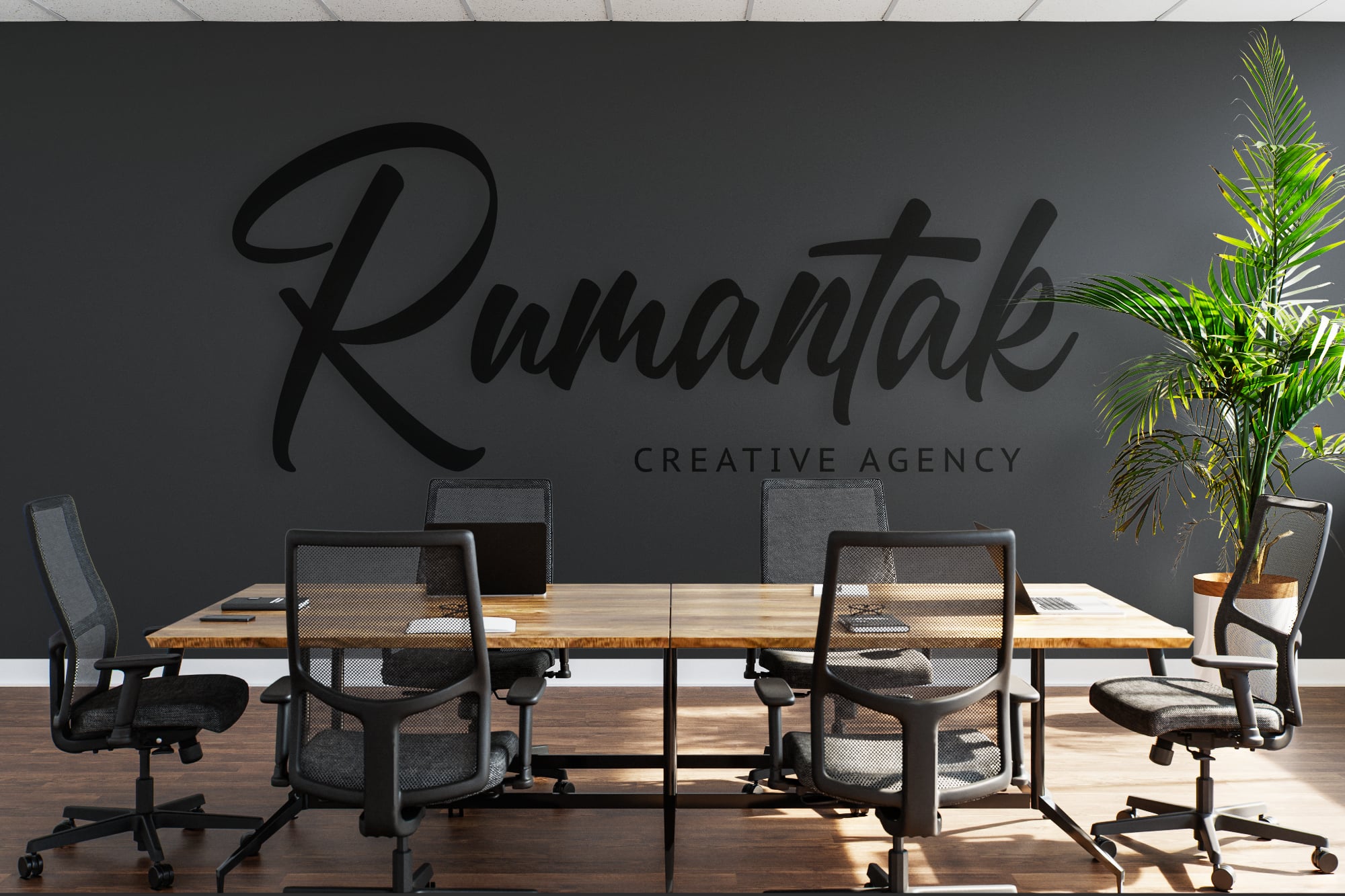 Modern office interior with wooden desk, mesh chairs, potted plant, wall decal mockup displaying "Rumantak Creative Agency" branding on dark gray wall
