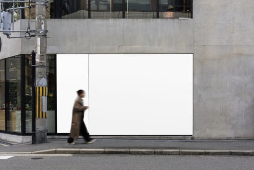 Blank white billboard on urban storefront with blurred person walking past concrete building