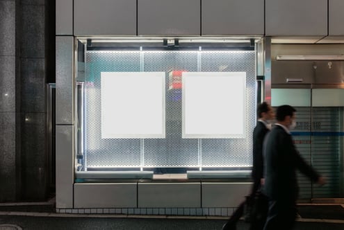Storefront window mockup with two blank poster frames on perforated metal backdrop, urban street scene with blurred pedestrians passing by