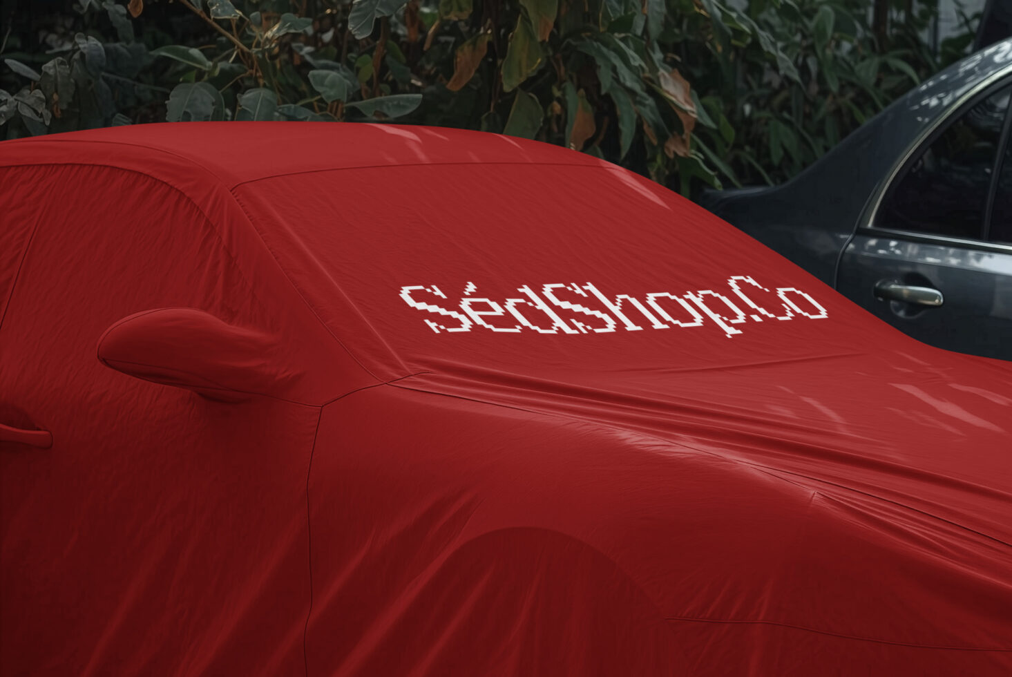 Bright red car cover with pixelated white text logo on hood, parked outdoors with greenery behind