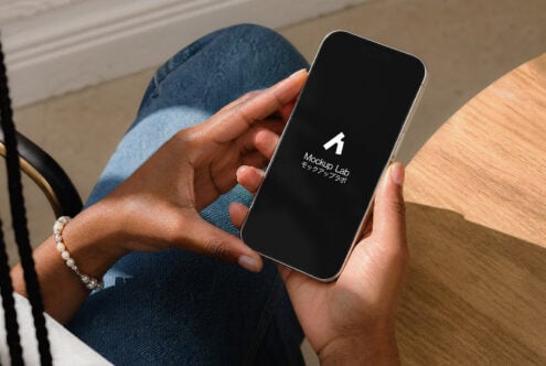 Person holding phone with black screen showing Mockup Lab logo, sitting at wooden table wearing jeans
