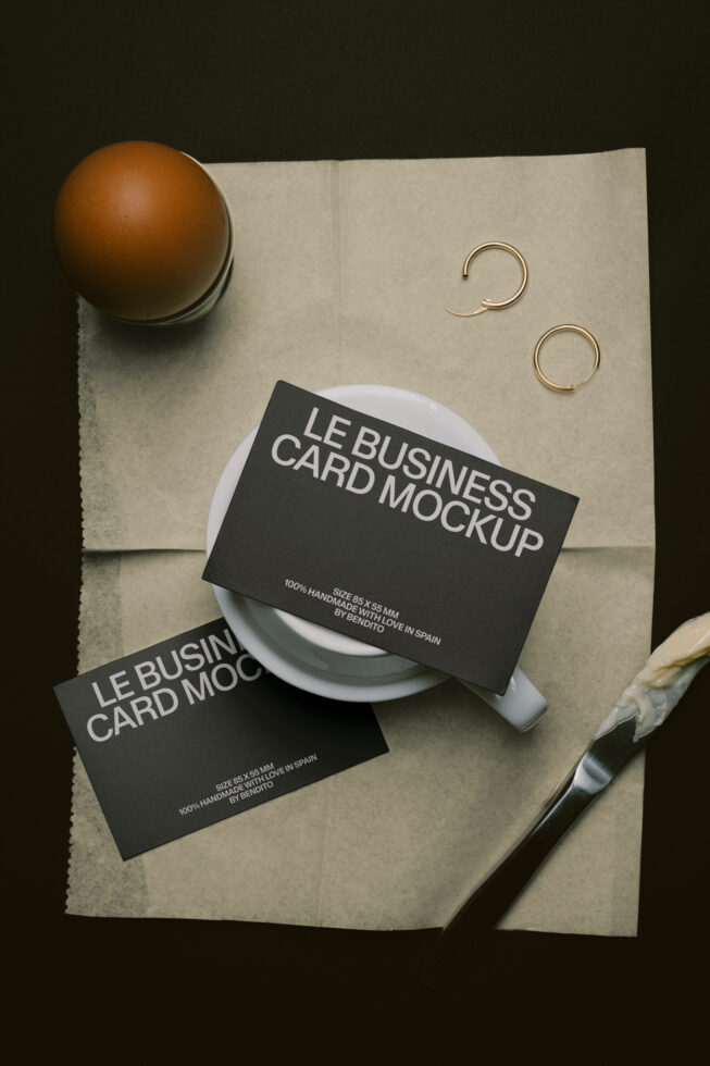 Two black business cards styled on cream paper with ceramic cup, bronze sphere, gold rings, plus brush