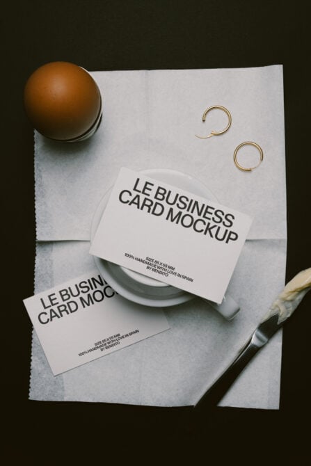 Minimalist flat lay showing two business cards on textured paper with ceramic bowl, brass rings, paintbrush