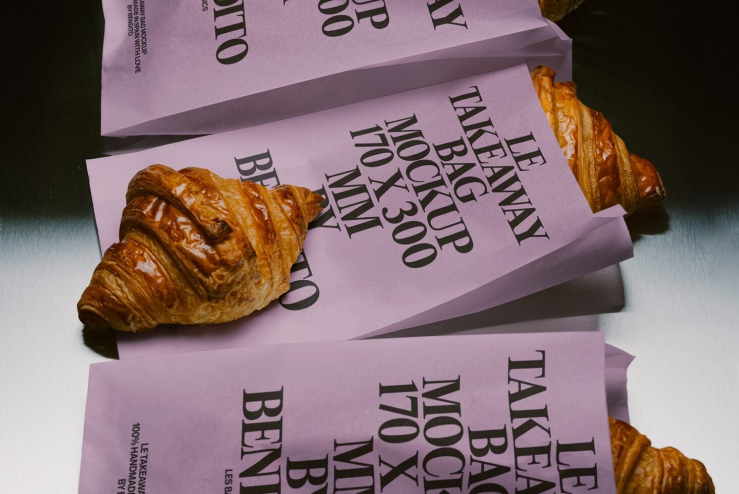 Stacked pink box mockups with bold black serif text, fresh croissants scattered around on white surface
