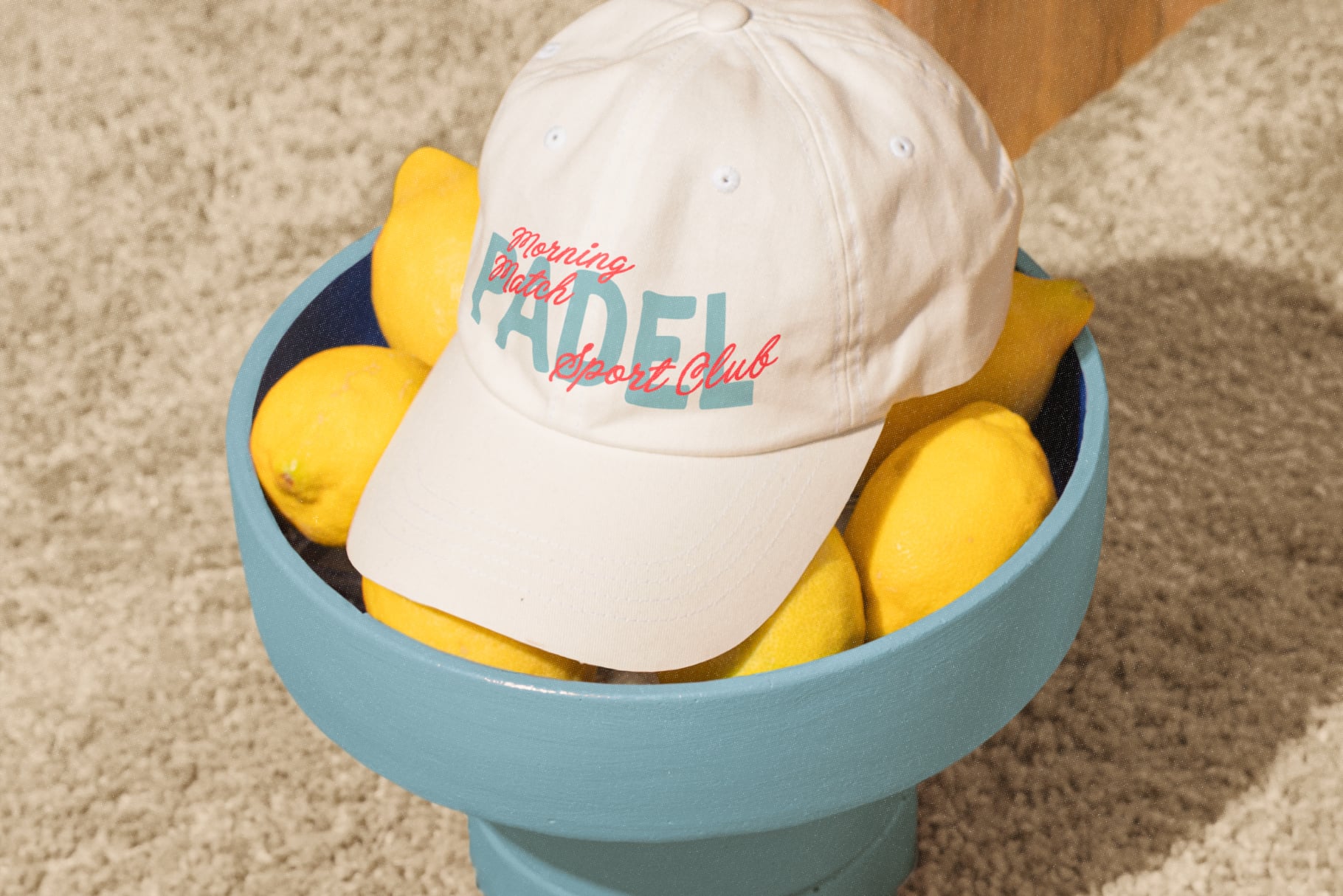 Cream baseball cap with retro script logo sitting in blue bowl filled with bright yellow lemons on sandy background