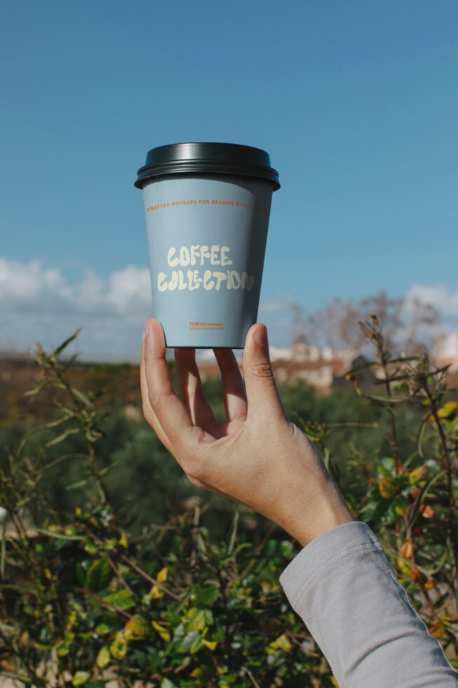 Hand holding coffee cup mockup against blue sky with field backdrop