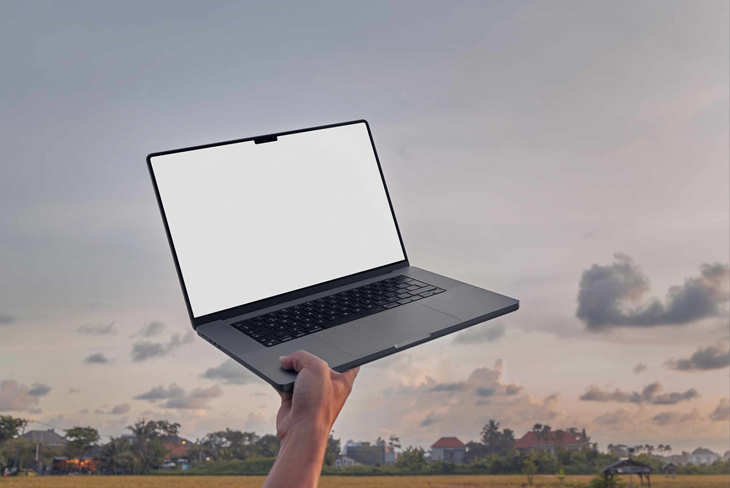 Hand holding dark laptop with blank screen against sunset sky over rural landscape