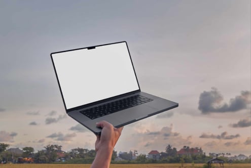 Hand holding dark laptop with blank screen against sunset sky over rural landscape