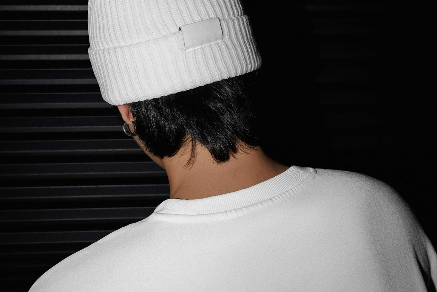 Person wearing white beanie with blank label tag, shot from behind against dark striped background