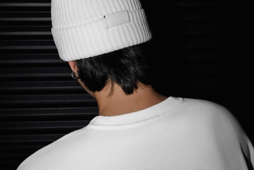 Person wearing white beanie with blank label tag, shot from behind against dark striped background