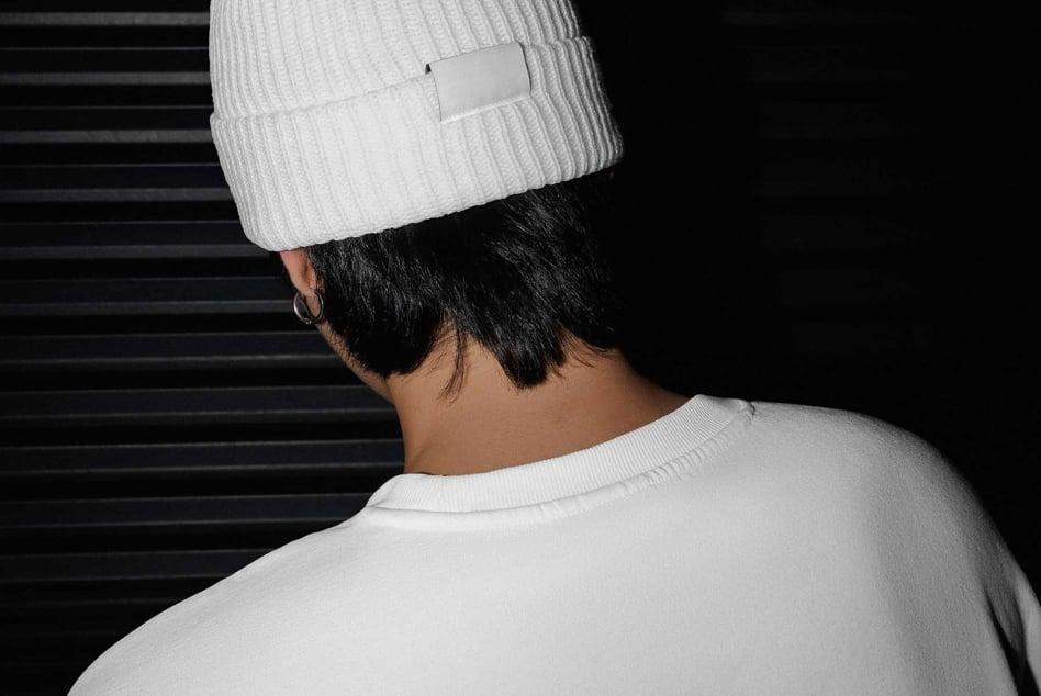 Person wearing white beanie with blank label tag, shot from behind against dark striped background