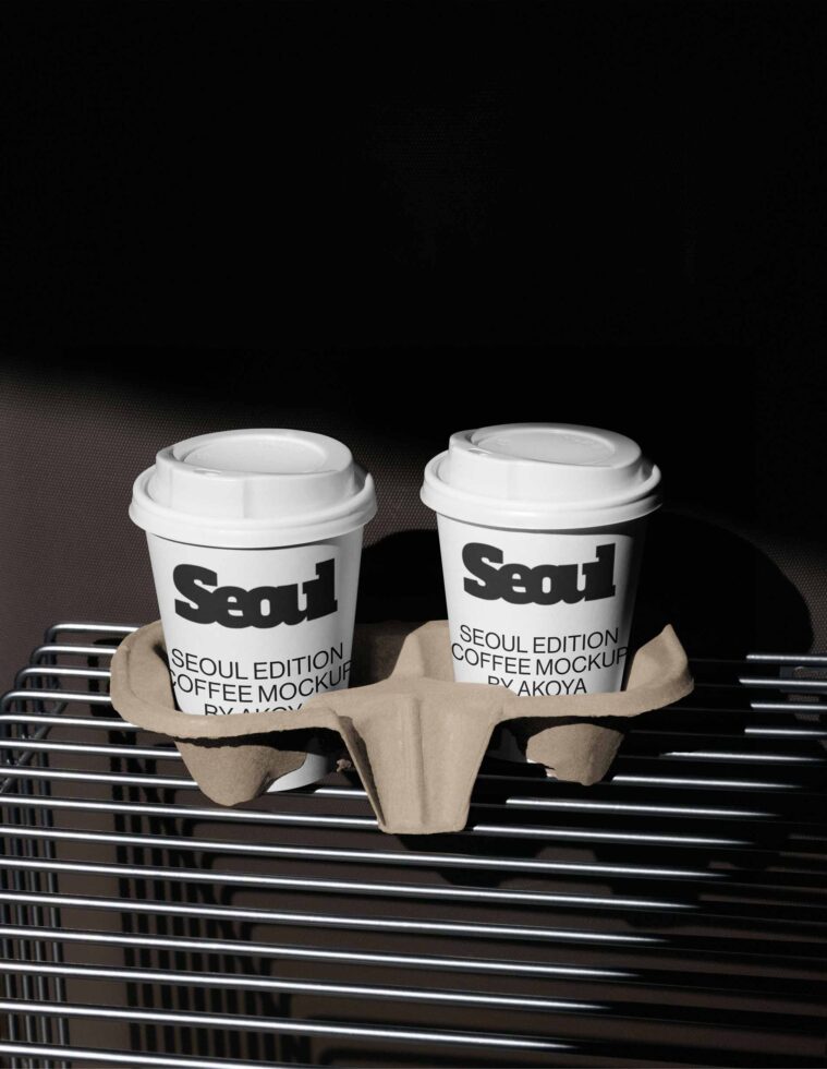 Two white coffee cups with Seoul branding sitting in cardboard holder on striped surface