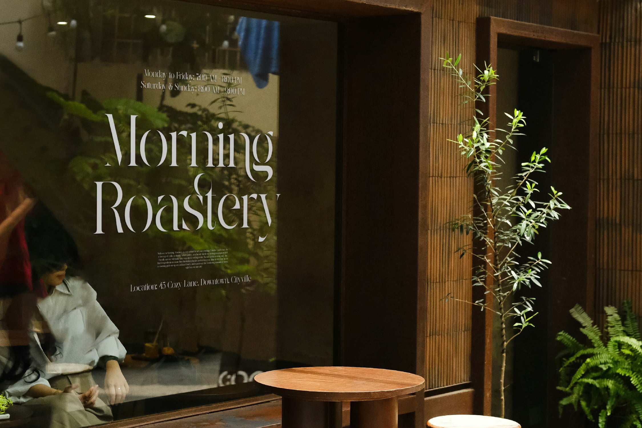 Storefront window for Morning Roastery coffee shop with elegant serif typography, wood table, greenery, hours posted