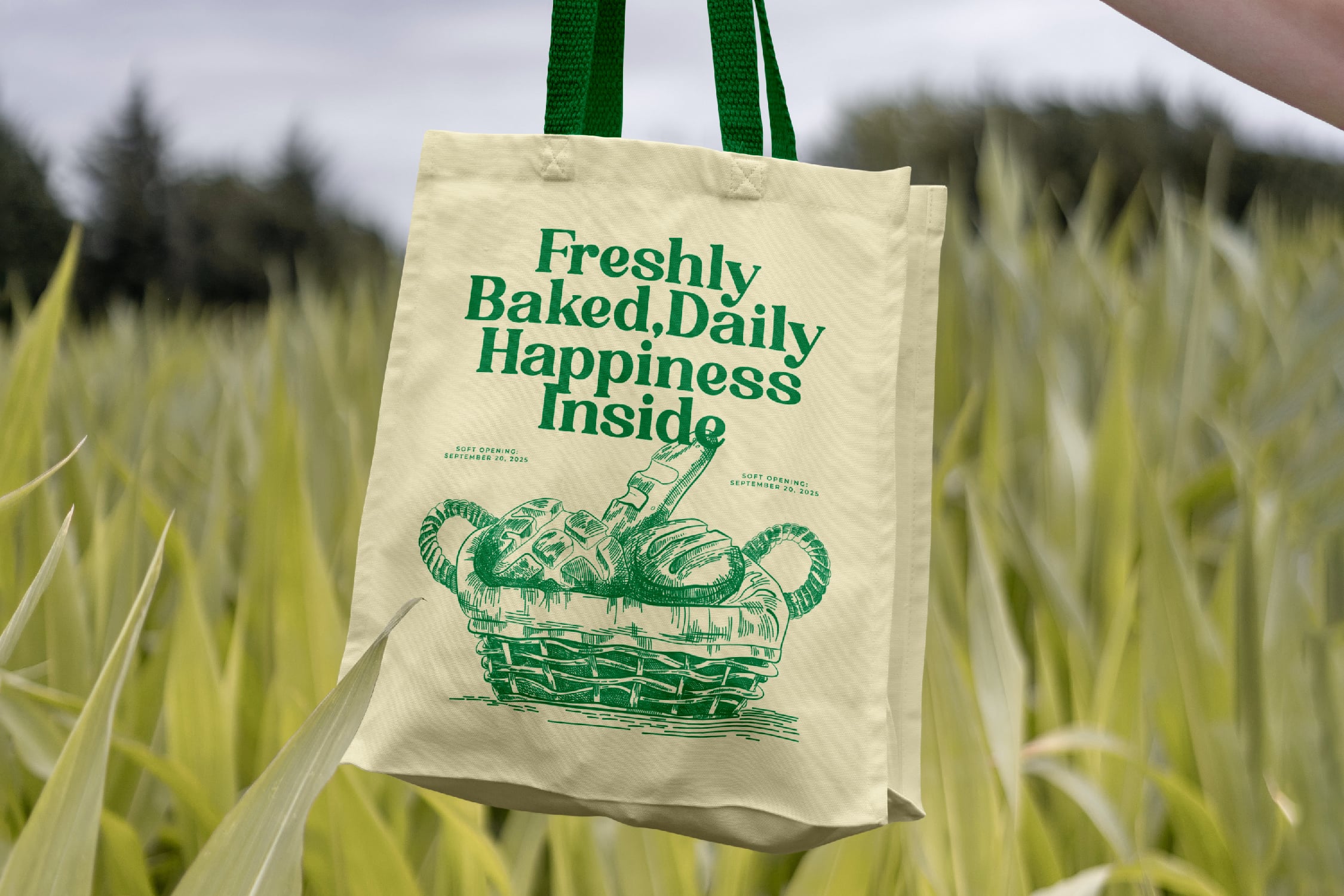 Cream tote bag with green bread basket illustration held in wheat field