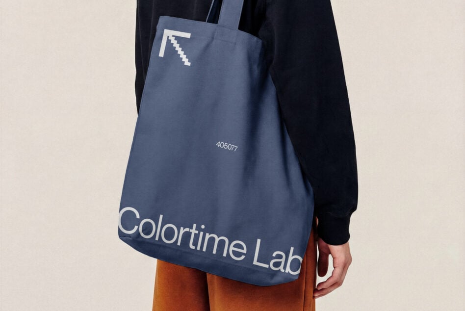 Blue tote bag mockup with pixel logo, worn over shoulder with navy jacket, orange pants visible