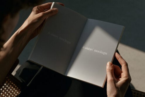 Hands holding open hardcover book mockup with natural light casting shadows across pages